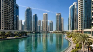 Dubai Lake Towers skyline with modern high-rise buildings surrounding a turquoise waterfront, palm-lined promenade, and clear blue sky in Dubai Lake Towers district.