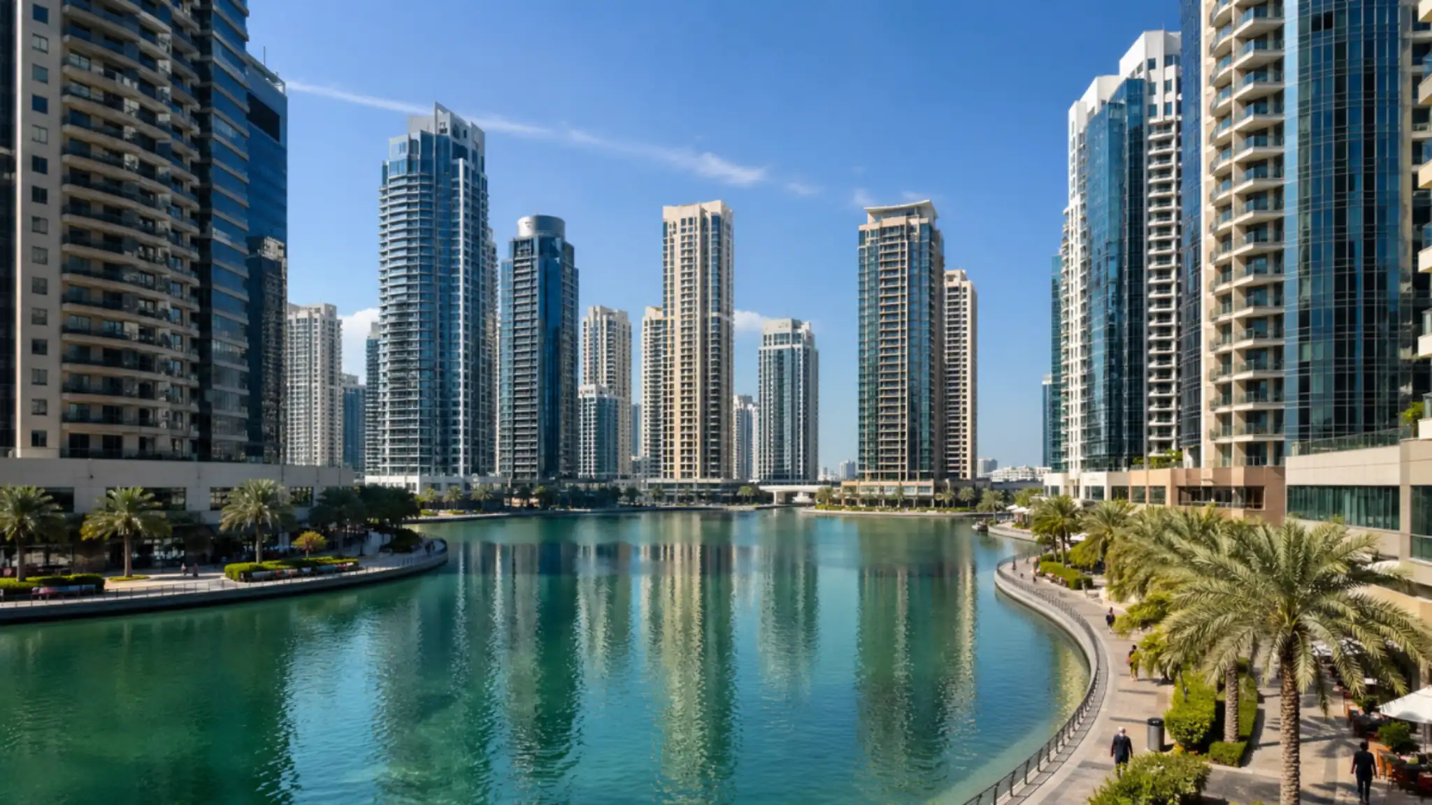 Dubai Lake Towers skyline with modern high-rise buildings surrounding a turquoise waterfront, palm-lined promenade, and clear blue sky in Dubai Lake Towers district.